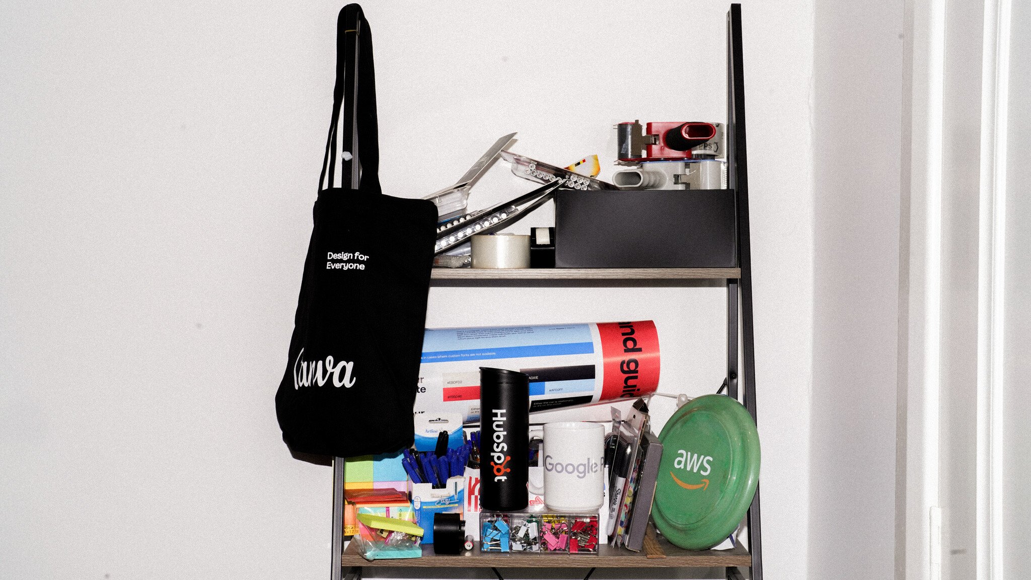 Tech company merchandise display featuring partner brands: a Canva tote bag, HubSpot water bottle, Google mug, and AWS frisbee. Additional marketing materials arranged on wooden shelves against a white wall.