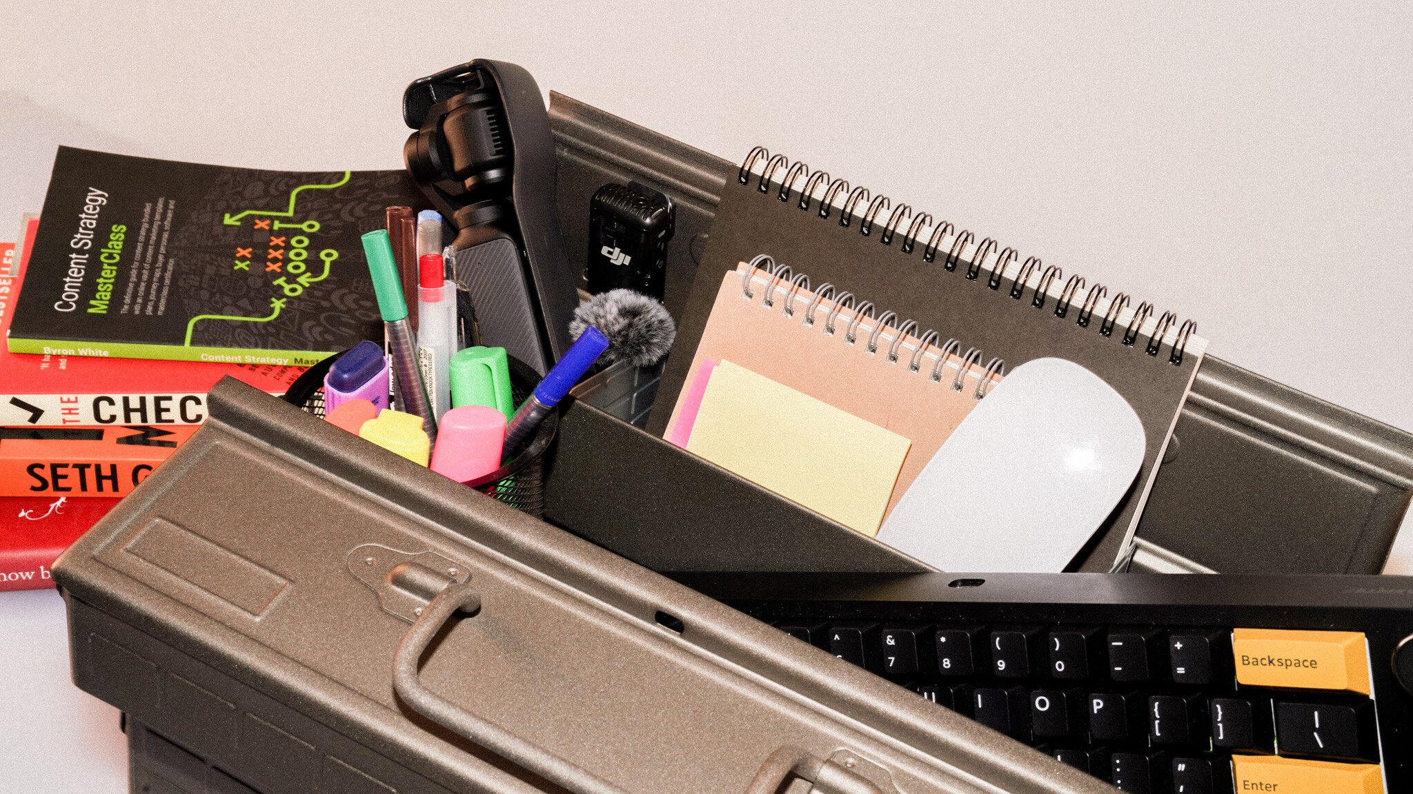 A desktop workspace toolkit including a DJI microphone, colorful highlighters, spiral notebooks, sticky notes, a computer mouse, keyboard with custom keycaps, and marketing strategy books. The items are organized in a desk organizer, suggesting a content creator or marketer's essential tools.