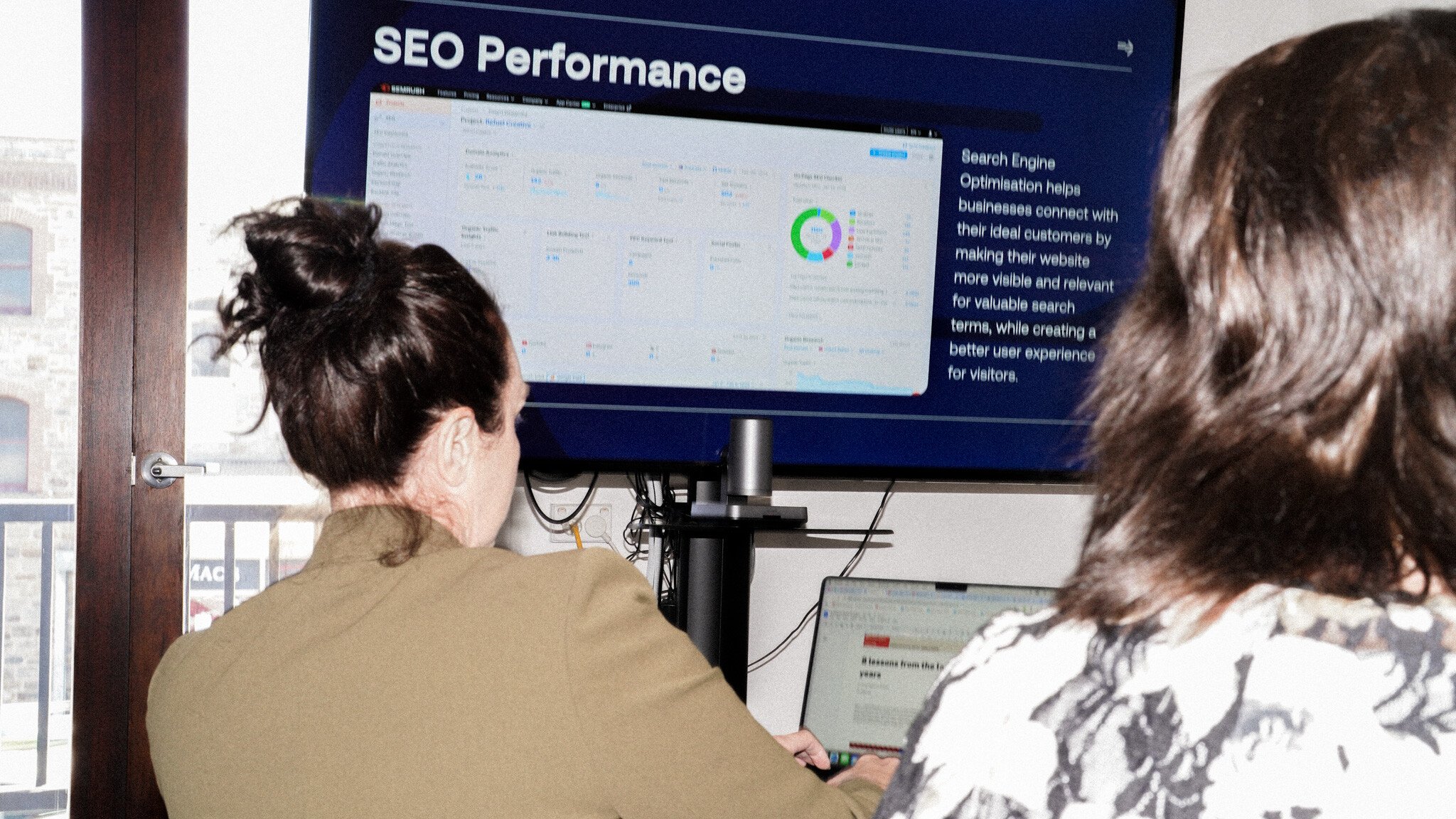 Two people looking at a large screen displaying SEO Performance analytics with a circular chart. They're viewing data on laptops while sitting at a desk below the mounted monitor.