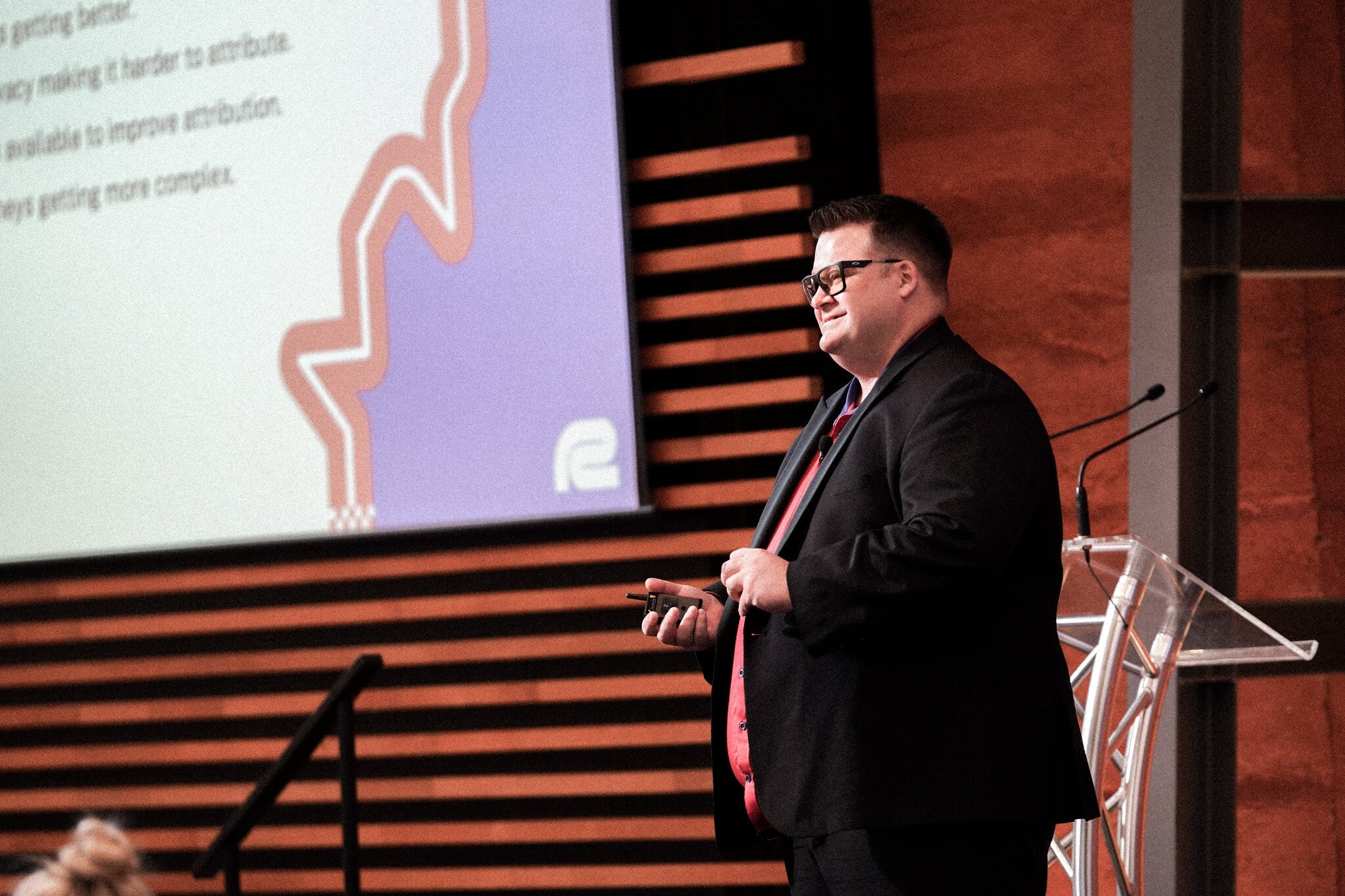 Ryan Jones presenting at a conference, wearing a black suit and red shirt, standing at a podium with a Refuel Creative presentation slide visible behind him. The warm-toned backdrop features wooden panels, and the presentation screen shows text and a stylised figure graphic.