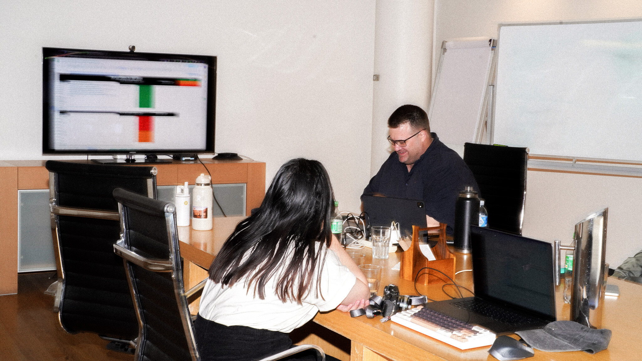 Two people in an office setting working at a desk, with a monitor displaying data visualisation in the background. The workspace includes laptops, water bottles, and a whiteboard visible in the corner. The setting appears to be a collaborative meeting or work session