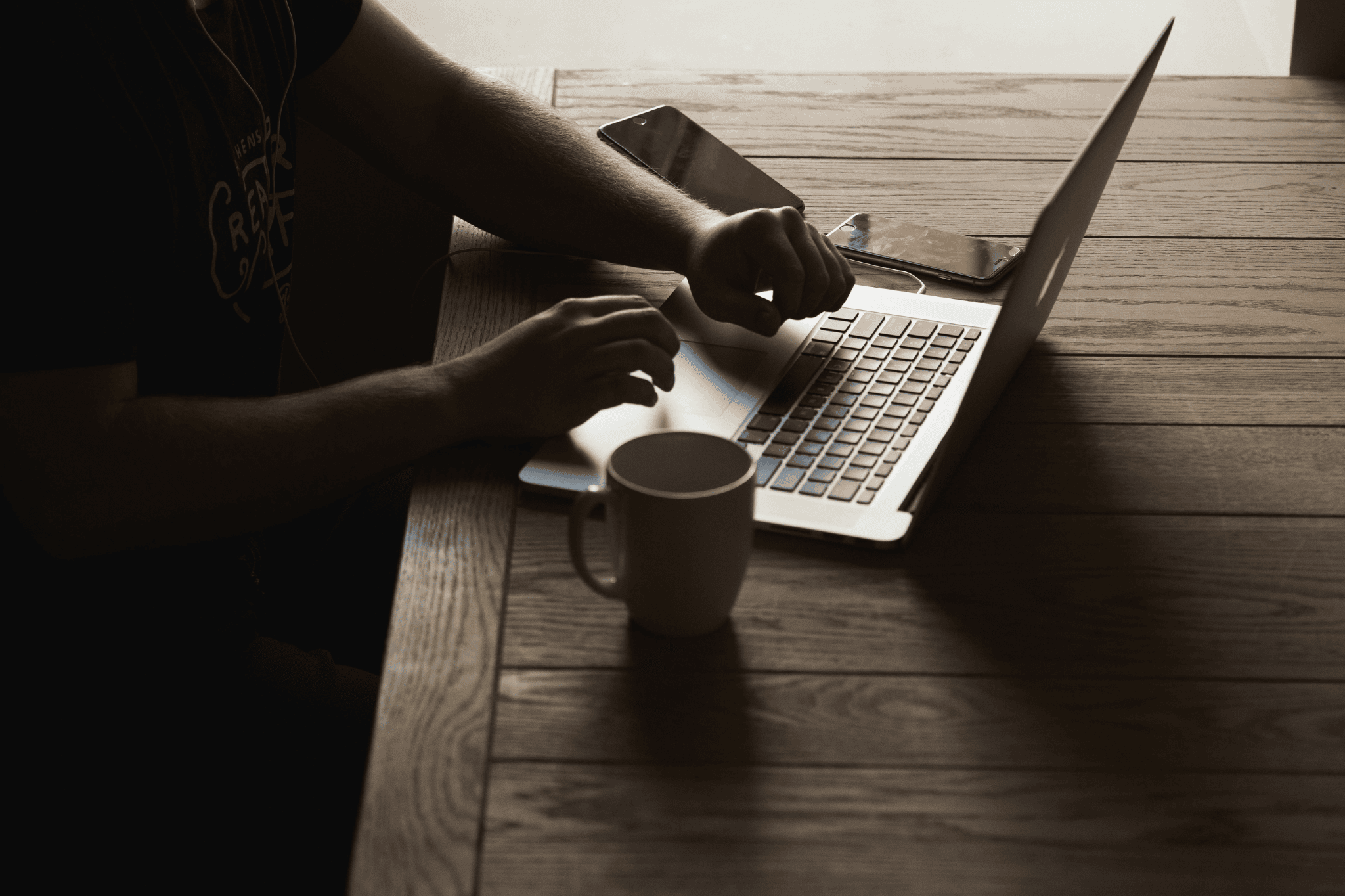 man at table typing on a laptop with a coffee mug and mobile devices nearby 